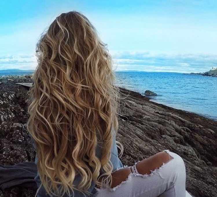 Woman with long beachy wavy hair sitting by the ocean, summer hair care